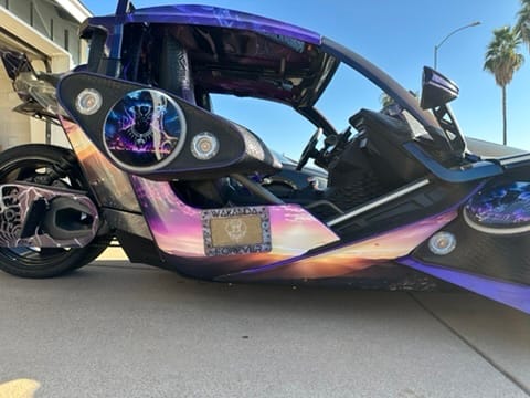 Custom purple-pink three-wheeled sports vehicle with open cockpit, parked on a driveway beside a house.