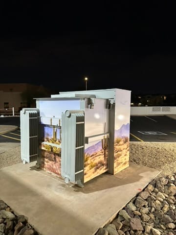 Outdoor electrical transformer wrapped in a nature landscape mural, panels opened to reveal equipment, at night in a parking lot.”,