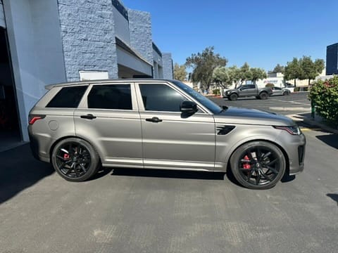Side view of a gray SUV with black wheels and red brake calipers parked in a sunny lot beside a blue-and-white building.