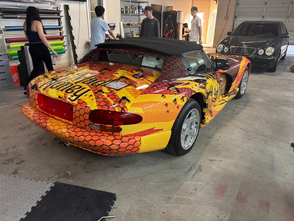 Bright yellow–orange wrap with a honeycomb pattern on a convertible sportscar in a workshop, with people and a black car in the background.