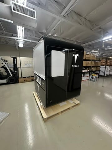 Black Tesla-branded vehicle cabin mock-up on a wooden pallet in a warehouse, with shelves and equipment in the background.