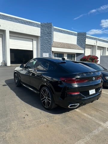 Black luxury coupe parked in a lot in front of a row of white/gray industrial buildings with roll-up doors; sun reflects off its glossy surface, rear three-quarter view.