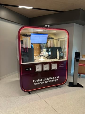 Red-framed self-serve coffee kiosk with a top screen and labeled dispensers; a worker is visible behind the glass enclosure.