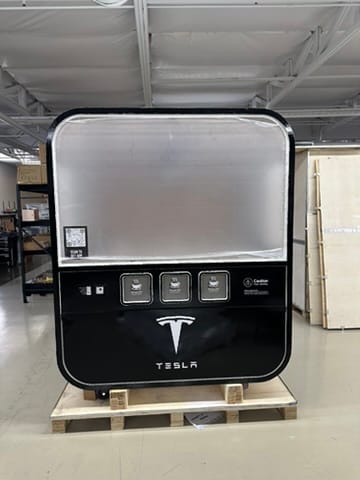 Tesla home charging unit on a wooden pallet in a warehouse, black enclosure with Tesla logo and three access panels.