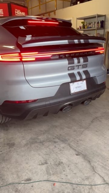 Rear view of a silver Porsche GTS SUV in a workshop, with a continuous red taillight strip and quad exhausts visible at the rear coolerside.