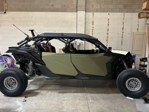 Side view of a two-seat off-road dune buggy with olive-green panels, black frame and exposed suspension, parked in a warehouse.