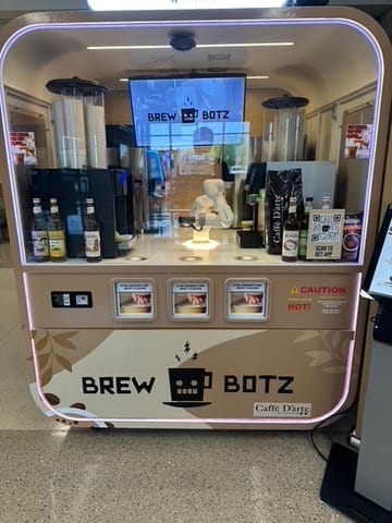 Self-service beverage vending kiosk labeled Brew Botz with a glass enclosure and a robotic-looking attendant behind the counter.