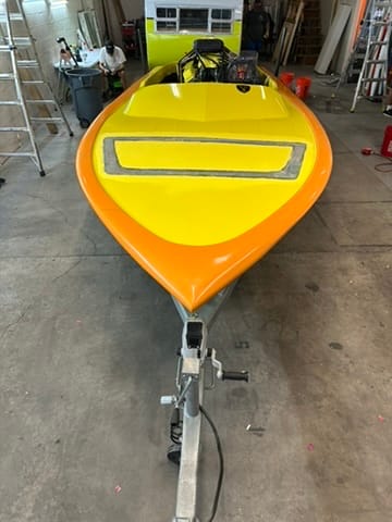 Bright yellow and orange speedboat on a metal trailer inside a workshop, facing away from the camera.