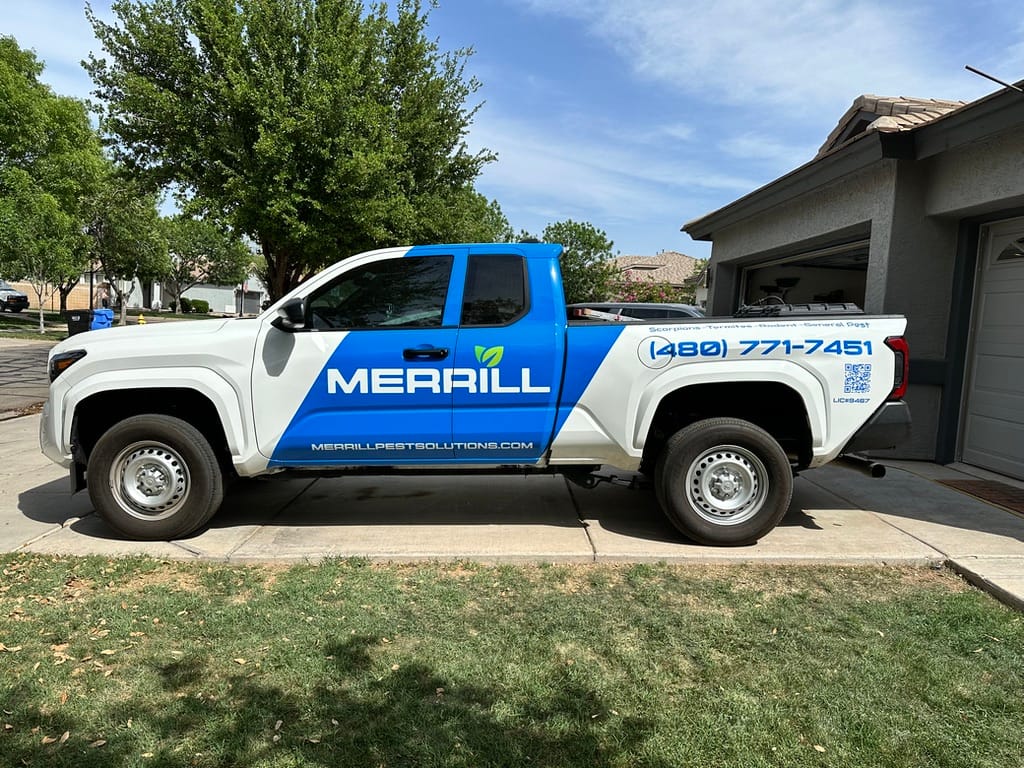 White pickup truck with blue Merril Pest Solutions branding parked on a suburban driveway near a garage.