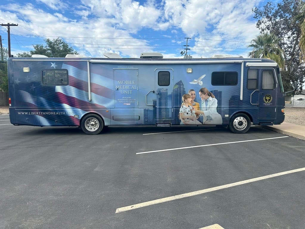 Blue mobile medical unit RV with American flag graphics parked in a lot; a doctor and nurse examine a child on the side of the bus.