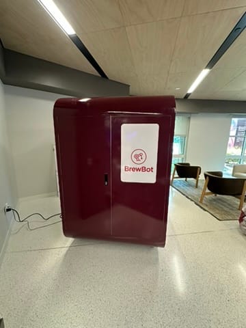 Maroon BrewBot automated beverage dispenser cabinet in a modern lounge with seating.