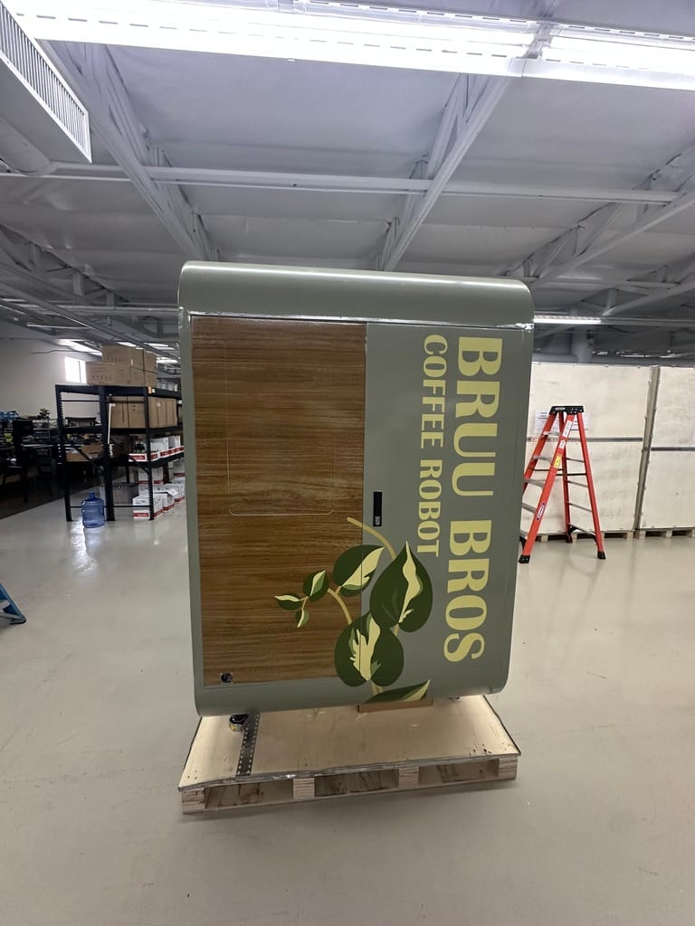 Gray coffee robot cabinet branded 'BRU BROS COFFEE ROBOT' with a wood panel and green plant decal, on a pallet in a warehouse setting.