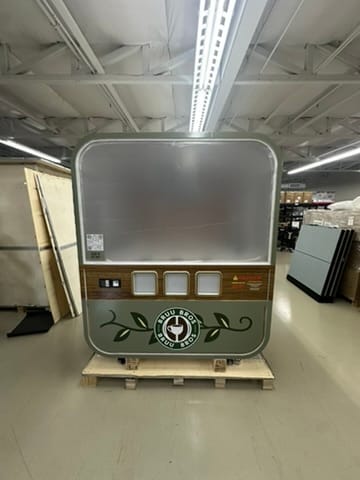 Front view of a retro-style beverage dispenser on a wooden pallet in a warehouse, with three small buttons and a green circular logo at the bottom.