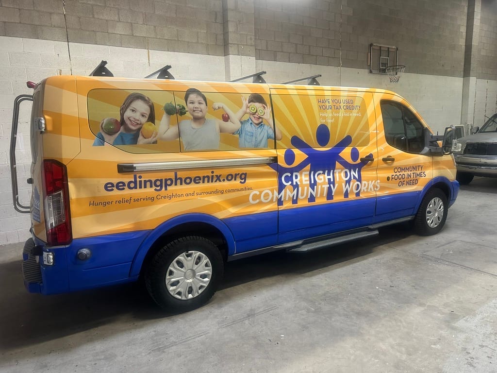 Branded orange–blue community service van for Creighton Community Works, promoting hunger relief and food donations inside a warehouse.