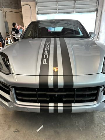 Front view of a silver Porsche SUV with dark racing stripes and Porsche emblem in a garage.